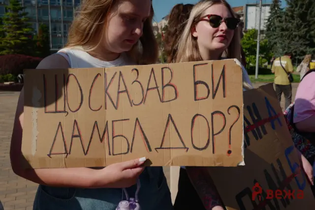 Two young people hold a cardboard protest sign with handwritten Cyrillic text at a sunny outdoor demonstration.