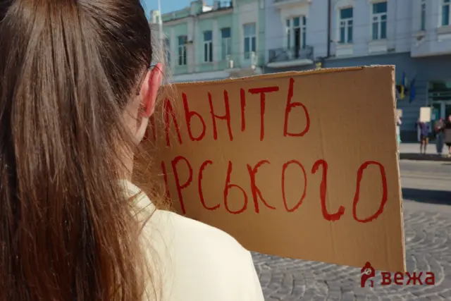 Back view of a person with long brown hair holding a cardboard sign with red handwritten Cyrillic text, on a city street backdrop.