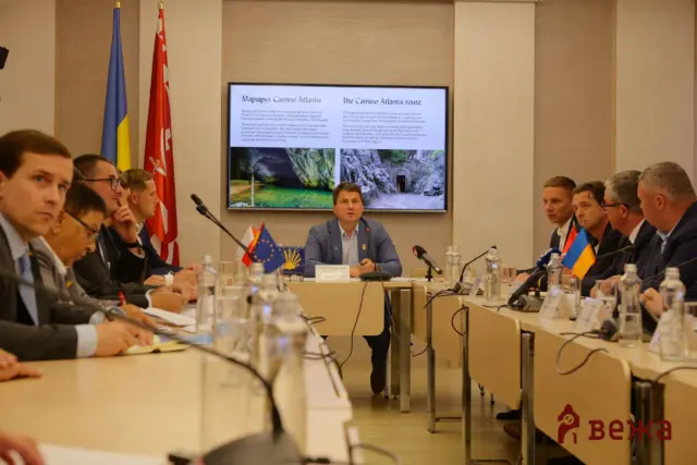 Conference room meeting with officials around a long table; a man speaks at the center with a large screen behind him.
