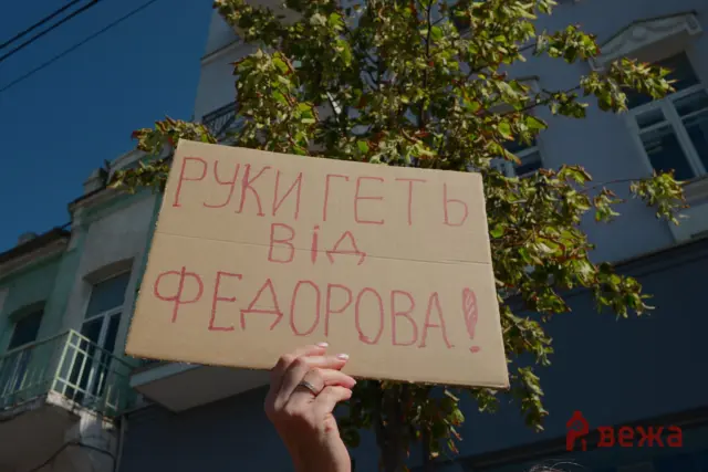Protester holds a cardboard sign with handwritten red Cyrillic text outdoors, blue sky and building in the background.