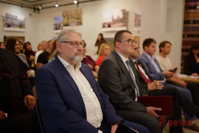 Audience at a formal event: foreground man in a navy suit and glasses, seated among attendees in a gallery-like room.