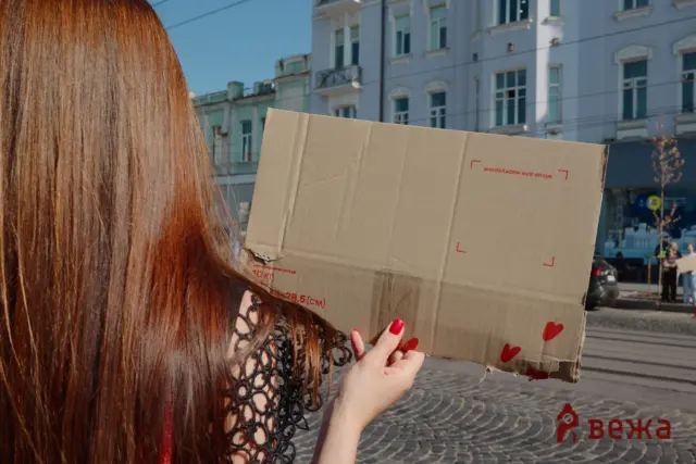 Person with long red hair holding a large cardboard box with red heart graphics outdoors on a sunny city street behind them.