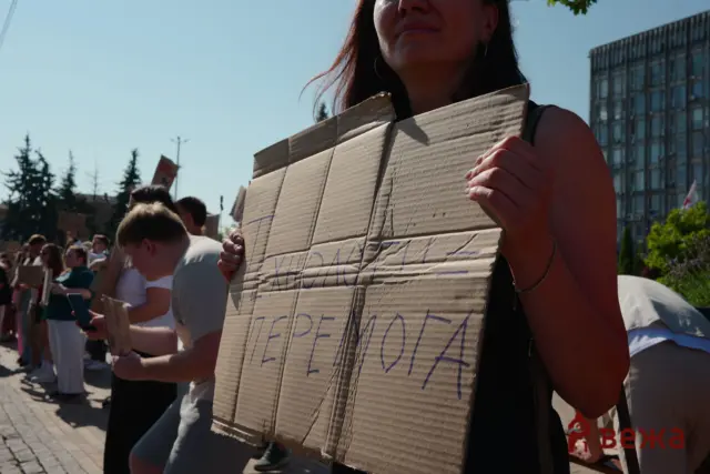 Protesters lined up along a city street, people holding cardboard signs; a foreground figure grips a large cardboard placard with handwritten text partially visible.