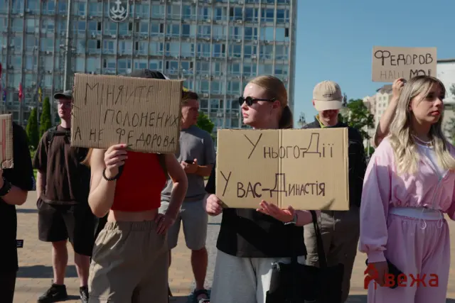 Group of protesters holding handwritten cardboard signs in a sunny urban square, with a glass-fronted building in the background.