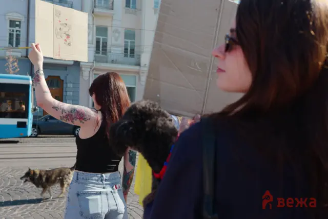 People on a sunny city street, one person raises a cardboard sign while a dog walks by; visible tattoos on the raised arm.