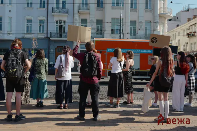 Group of people standing on a city street, some holding cardboard boxes overhead as if protesting or posing for a photo, with an orange vehicle and ornate buildings in the background.