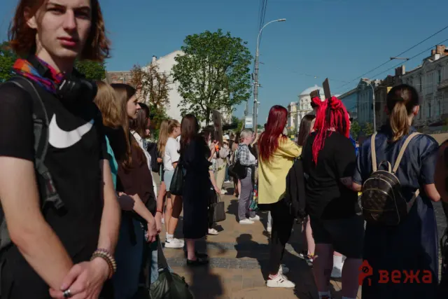 Crowd of people at a sunny street protest, holding placards with urban buildings and a tree in the background.