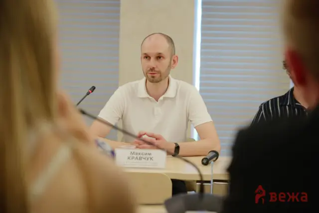Bearded man in a white polo speaking at a panel with microphones on the table, audience blurred in foreground.