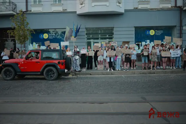 Crowd of protesters holding signs and Ukrainian flags on a city street, with a red Jeep in the foreground.