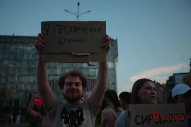 Protester raising a cardboard sign with handwritten Cyrillic text during an urban demonstration.