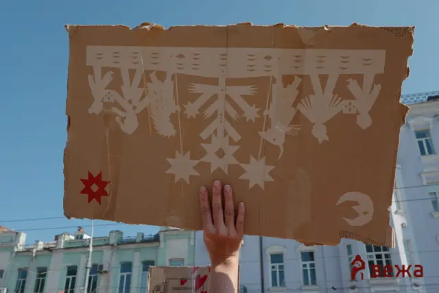 Person holding up a large cardboard sign with white geometric cutouts against a blue sky, city buildings in the background.