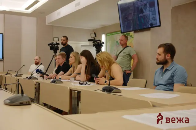 Panel of seven people seated at a conference table with microphones; two cameramen filming in a briefing room.
