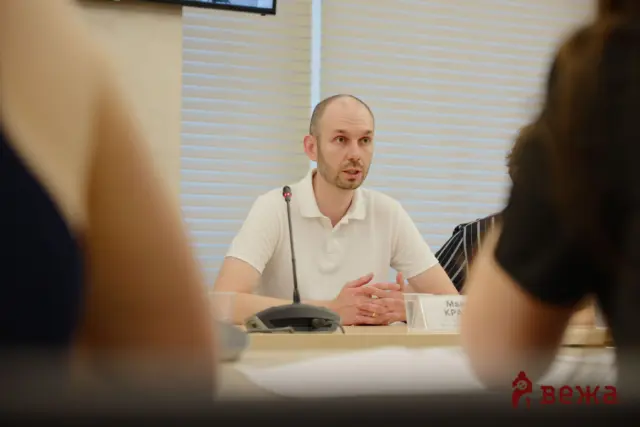 Man in a white polo shirt speaks into a microphone at a panel table during a meeting, with blurred attendees in the foreground.