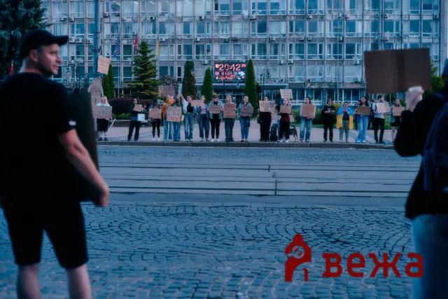 Group of protesters holding signs with Cyrillic text in a city square, visible in front of a modern office building; blurred people frame the sides of the scene.