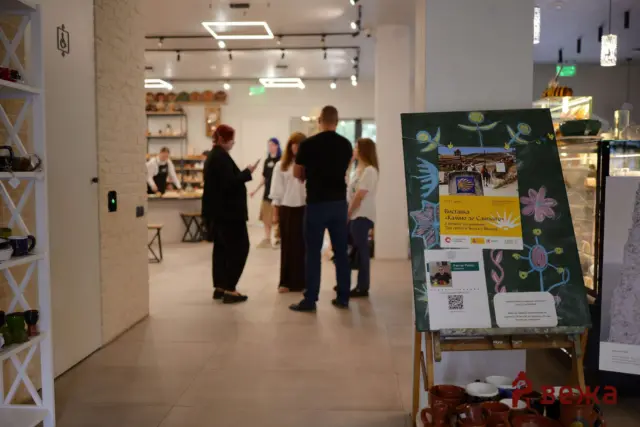 Indoor cafe scene with a group of people chatting near the counter; a colorful poster on an easel is in the foreground for viewing.