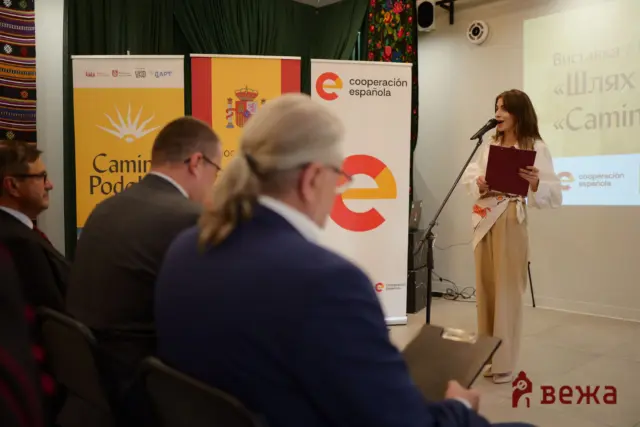 Female speaker at a podium delivering a presentation; banners behind her read 'cooperación española' with colorful designs, and an audience sits in foreground.