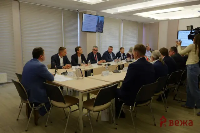 Panel of formally dressed professionals seated around a long conference table, with a camera operator recording on the right and screens on the walls.