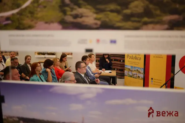 Group of people seated in a conference room, facing a speaker, with bright posters banners on the right side of the scene