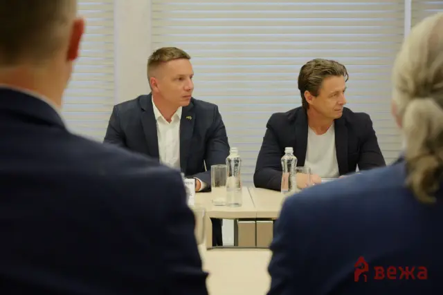 Two men in dark blazers seated at a conference table during a formal meeting, water bottles in front of them.