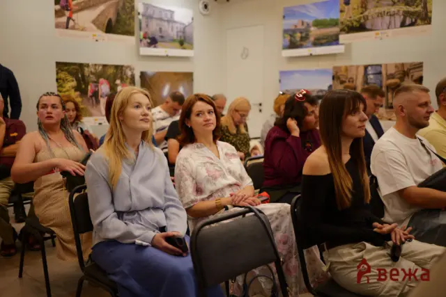 Group of adults seated in a classroom-like room, looking toward the front, with large travel photos on the walls.