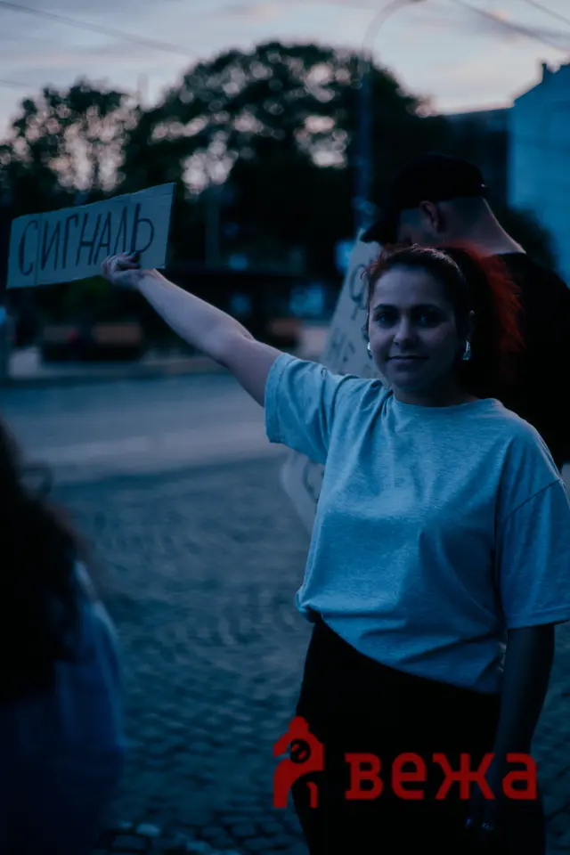 Young woman in a gray shirt stands outdoors at dusk, arm raised holding a cardboard protest sign with Cyrillic text partially visible behind her.