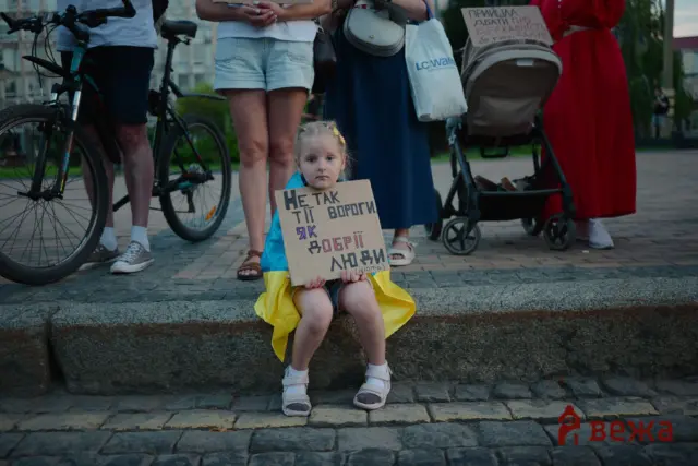 Young girl in a yellow dress sits on a stone curb holding a cardboard protest sign with handwritten Ukrainian text, while adults with bicycles stand behind her.