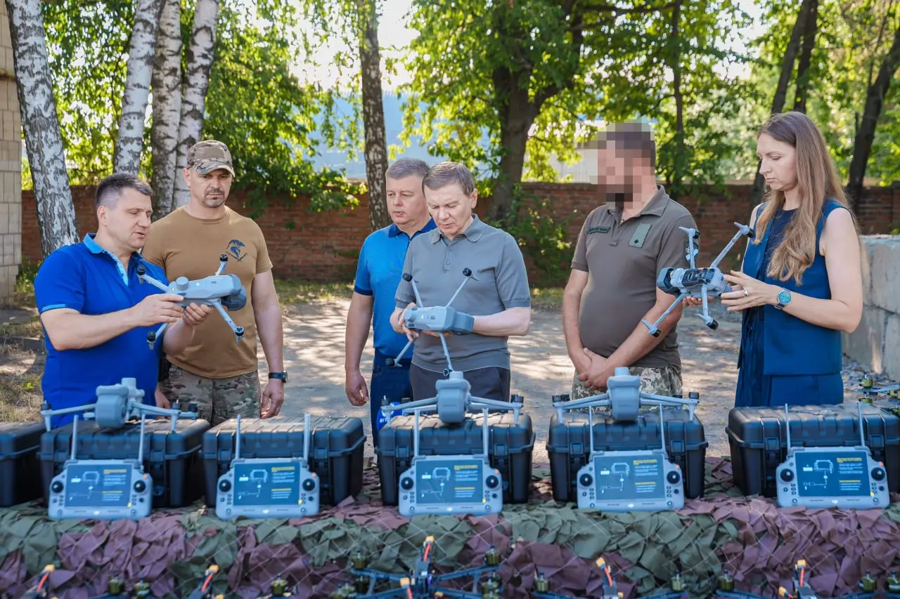 Group of six people outdoors examining drone controllers and equipment laid out on a table with protective cases and camouflage cloth, likely a training or demonstration session.