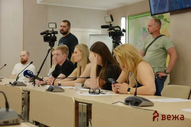 Panel of five people seated at a desk with microphones, two cameras filming behind them, and a map on the wall in a conference room.