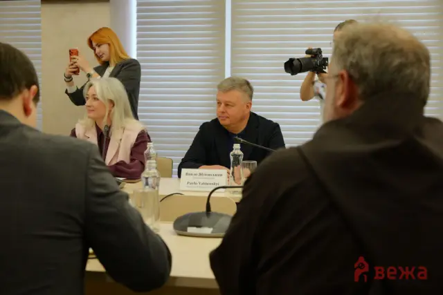 Meeting scene with a woman using a red phone to photograph the event while others sit at a conference table.