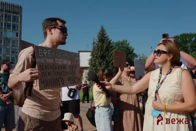 Man wearing sunglasses holds a cardboard protest sign with Cyrillic text as reporters and a crowd gather outdoors under a blue sky.