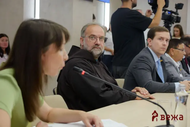 Bearded man with glasses sitting at a conference table and looking toward the camera, with a cameraman and other attendees in the background.