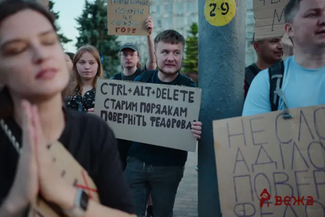 Group of protesters holding cardboard signs with Cyrillic text at a street demonstration; central sign reads 'CTRL+ALT+DELETE...'.