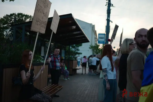 Group of people outdoors holding protest signs on sticks along a sidewalk, with a wooden planter and buildings in the background.