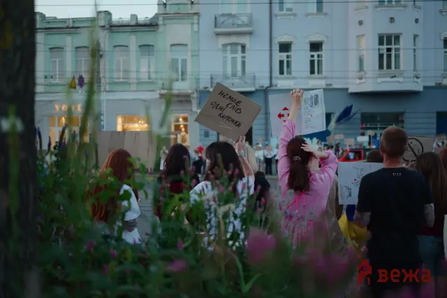 Crowd of protesters holding handmade signs on a city street with pastel buildings in the background, seen through plants in the foreground.