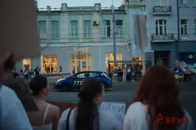 Crowd of protesters along a city street holding signs, a blue taxi driving past, and pastel storefronts in the background at dusk.