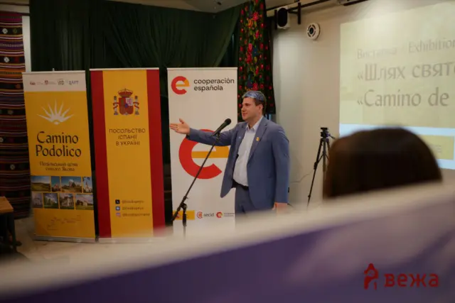 Man in a blue suit giving a presentation on a stage with Spanish banners nearby and a projector screen in the background.