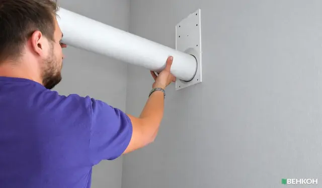 Man in a purple shirt inserting a white cylindrical duct through a wall mounting flange during installation (vent/air duct).