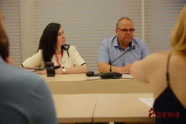 Two panelists seated at a conference table, woman on left and man on right, microphones in front of them during a meeting.