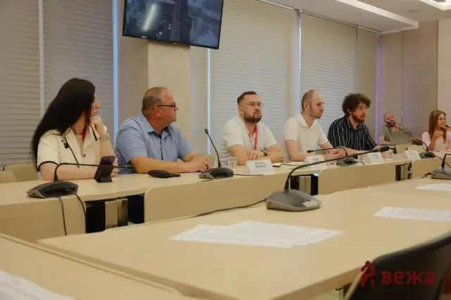 Panel of six people seated at a conference table with microphones and nameplates, in a meeting room with blinds and a wall-mounted screen above.