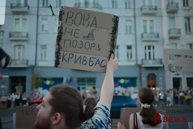 Protesters raise large cardboard signs with handwritten text in a street demonstration, urban buildings in the background.