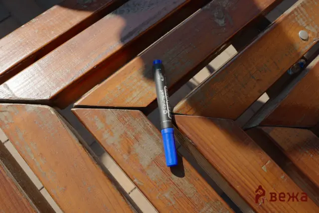 Blue marker pen resting on weathered wooden bench slats with sunlight shadows, and a logo in the lower corner.
