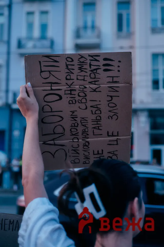 Person holding a cardboard protest sign with handwritten Cyrillic text, urban buildings in the background.