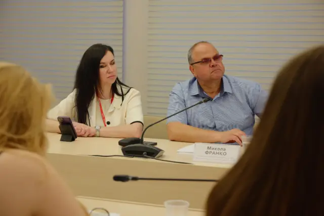 Two panelists sit at a conference table, a woman with long dark hair on the left and a man in glasses on the right, microphones in front of them.