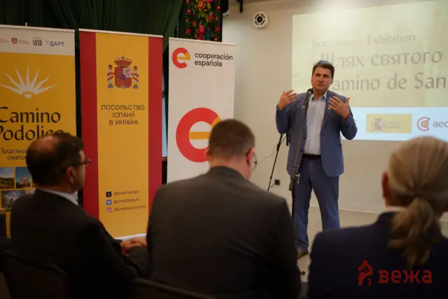 Speaker in a blue suit delivering a talk at a conference, microphone in hand, with Spanish-themed banners and a projection saying 'Exhibition' in the background.