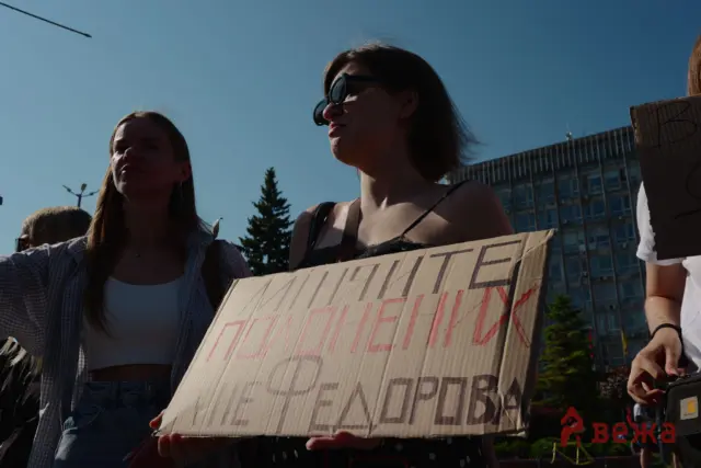 Group of people holding a large cardboard protest sign in an urban outdoor setting, with a building and blue sky in the background (faces shadowed by sun).