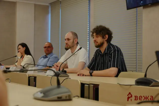 Panelists seated at a long conference table with microphones, discussing in a meeting room in front of blinds and screens.