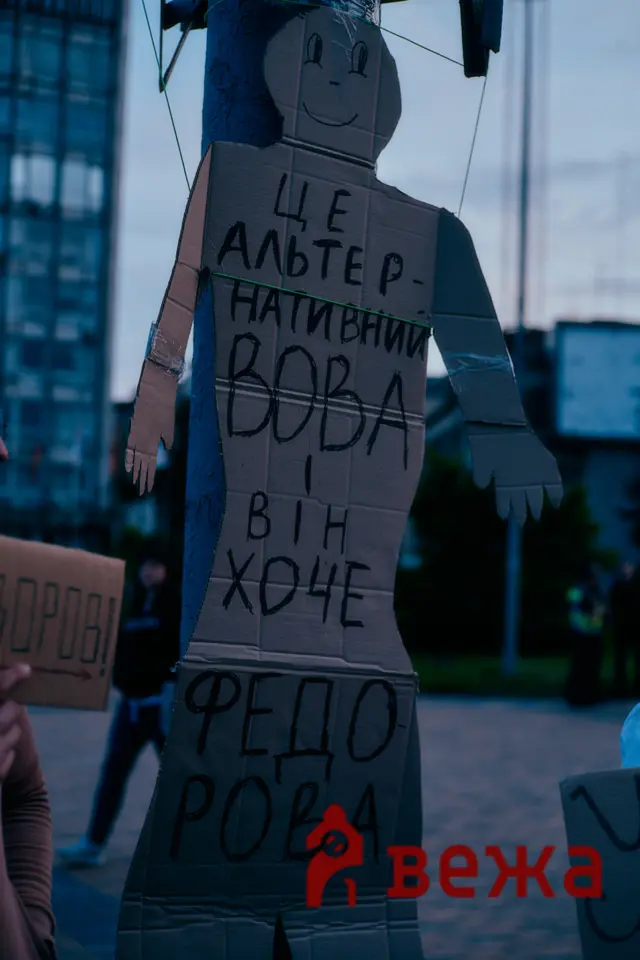 Cardboard cutout of a person with handwritten Cyrillic text on the torso, taped to a post in an urban outdoor setting, resembling a DIY protest figure.