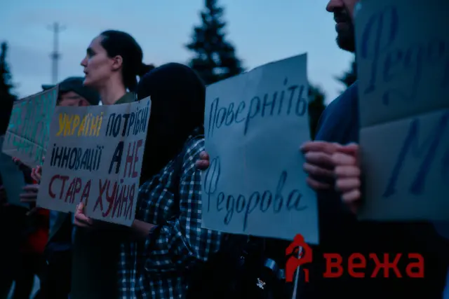 Group of protesters holding handwritten signs with Cyrillic text at dusk, facing forward in a crowd.