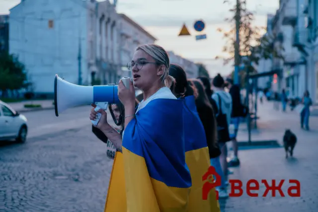 Woman wearing a blue and yellow flag speaks into a megaphone at a street protest with a line of people behind her.