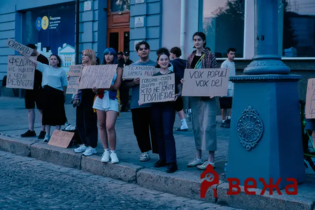 Group of young protesters standing on a city sidewalk, holding handwritten placards in Cyrillic. The blue-toned street scene shows a stone curb and buildings behind them.